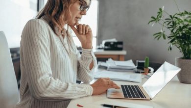 Woman in a striped blouse sits at a bright desk, typing on a laptop with papers nearby.