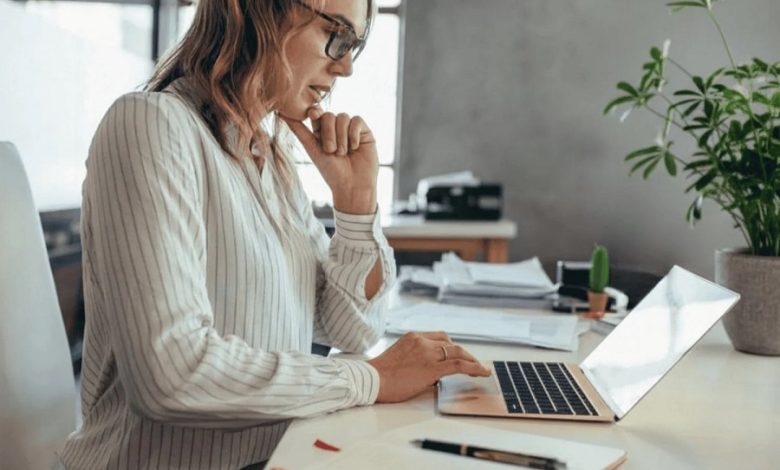 Woman in a striped blouse sits at a bright desk, typing on a laptop with papers nearby.