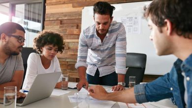Team of four collaborating around a table with documents and a laptop; one man stands and points at papers as others listen.