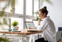 Woman with a bun and white blouse sits at a wooden desk near large windows, using a tablet with colorful charts while smiling.