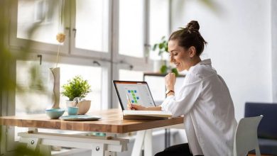 Woman with a bun and white blouse sits at a wooden desk near large windows, using a tablet with colorful charts while smiling.