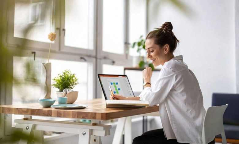 Woman with a bun and white blouse sits at a wooden desk near large windows, using a tablet with colorful charts while smiling.
