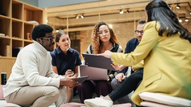 Five professionals of diverse backgrounds sit in a circle, reviewing a folder and notes in a bright office setting.