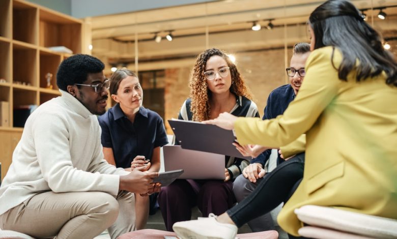 Five professionals of diverse backgrounds sit in a circle, reviewing a folder and notes in a bright office setting.